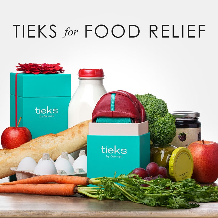 A pair of red Tieks flats in a teal Tieks box displayed among grocery items including a baguette, eggs, milk, broccoli, apples, grapes, carrots, and jars of preserves, symbolizing a food donation theme.